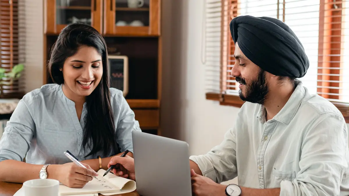 A smiling woman takes notes while a man in a turban works on a laptop beside her at a table at home