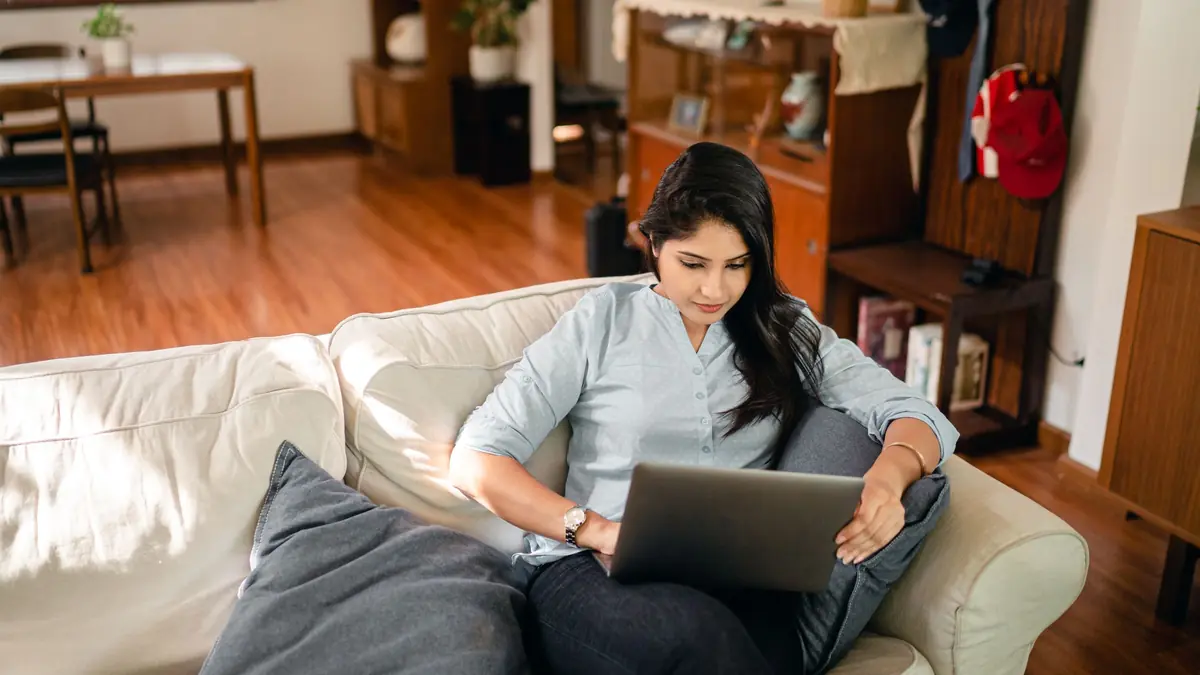 A young woman sits on a sofa at home working on a laptop