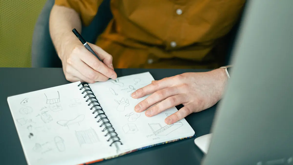 A hand sketches small drawings in a spiral notebook at a desk