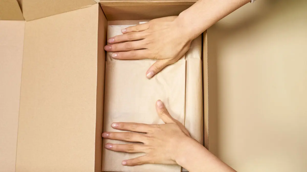 Two hands smooth kraft tissue paper inside an open cardboard shipping box
