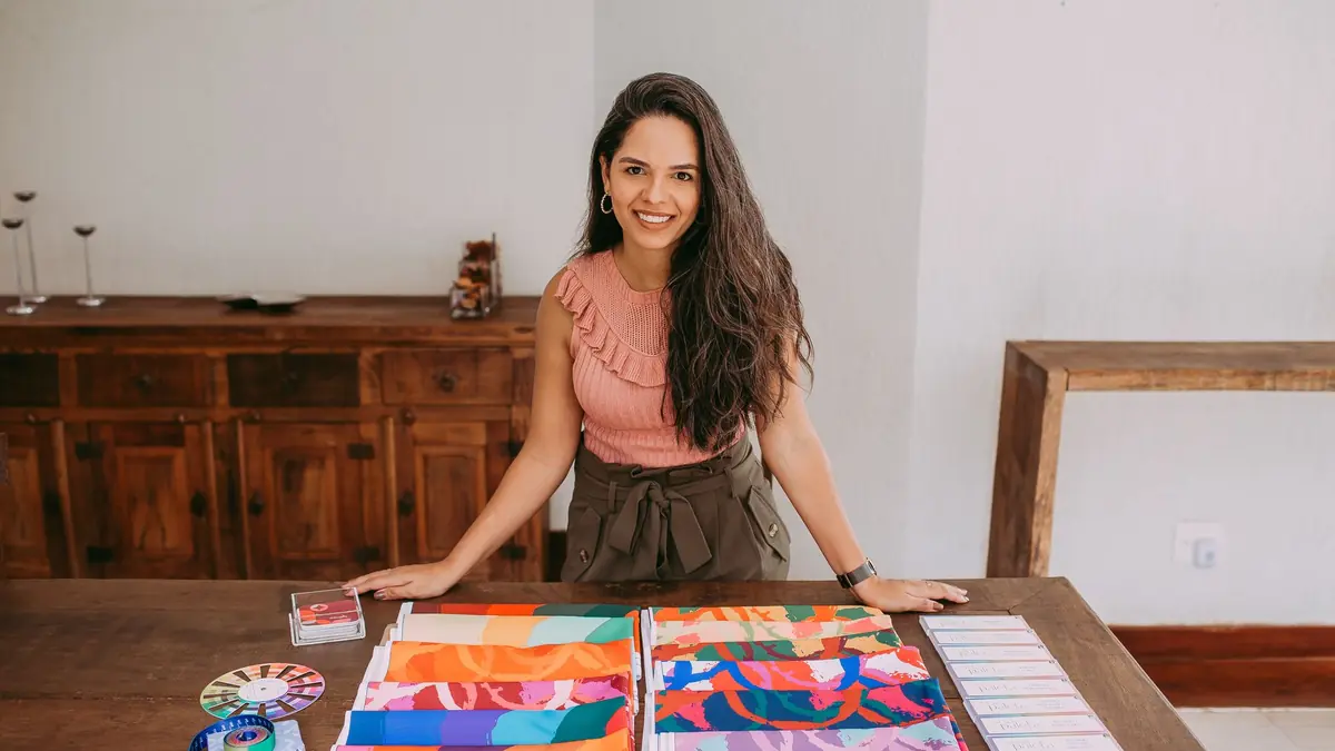 A smiling woman leans on a wooden table spread with colourful printed fabric samples and colour cards