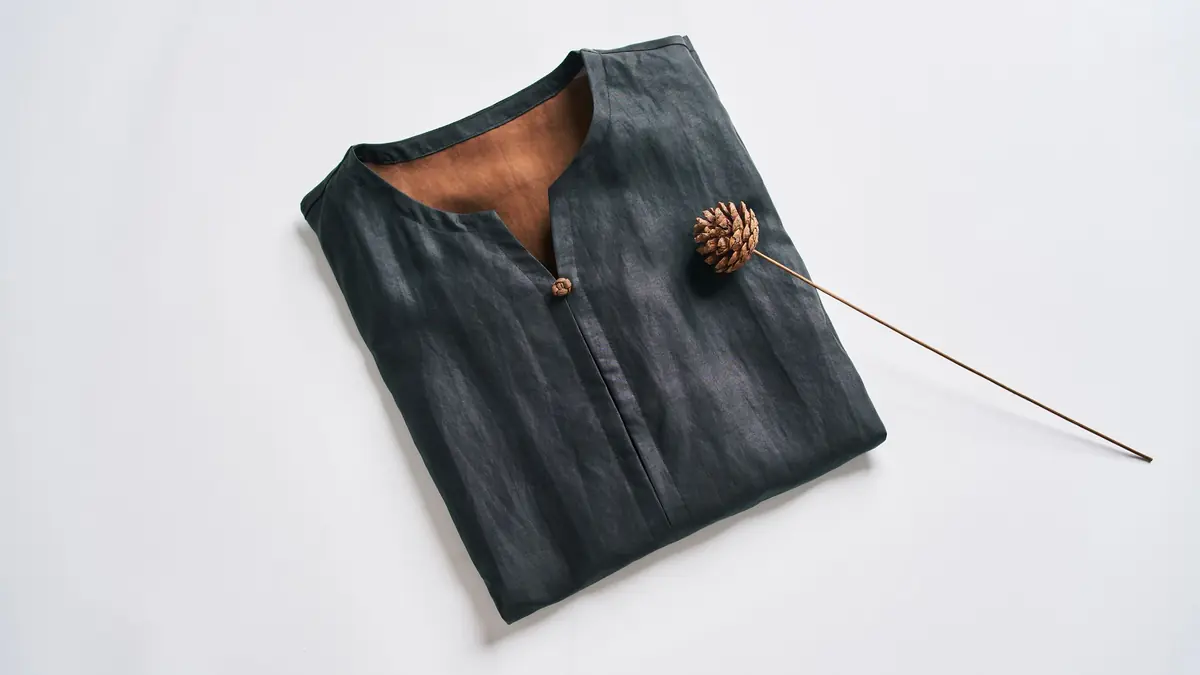 A folded dark linen kurta with a dried pine cone on a long stem, laid on a white background