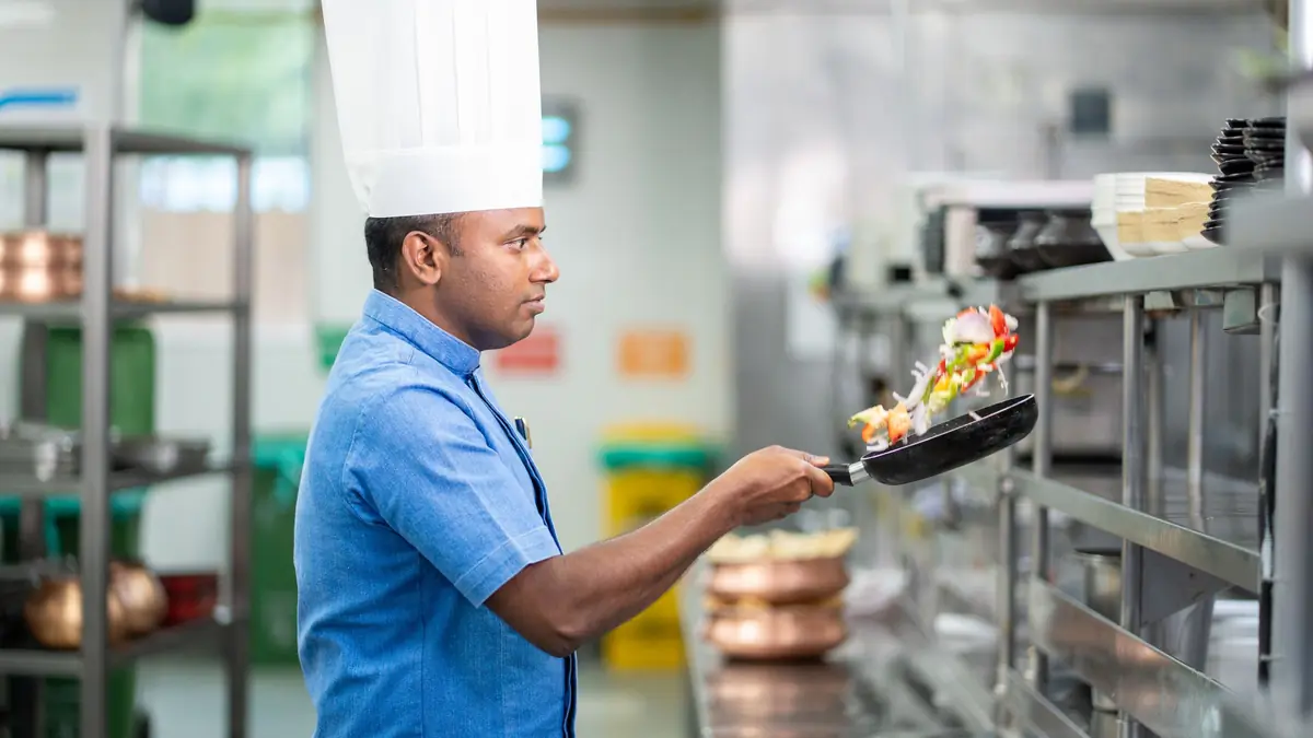 A chef in a white hat tosses vegetables in a pan in a steel commercial kitchen