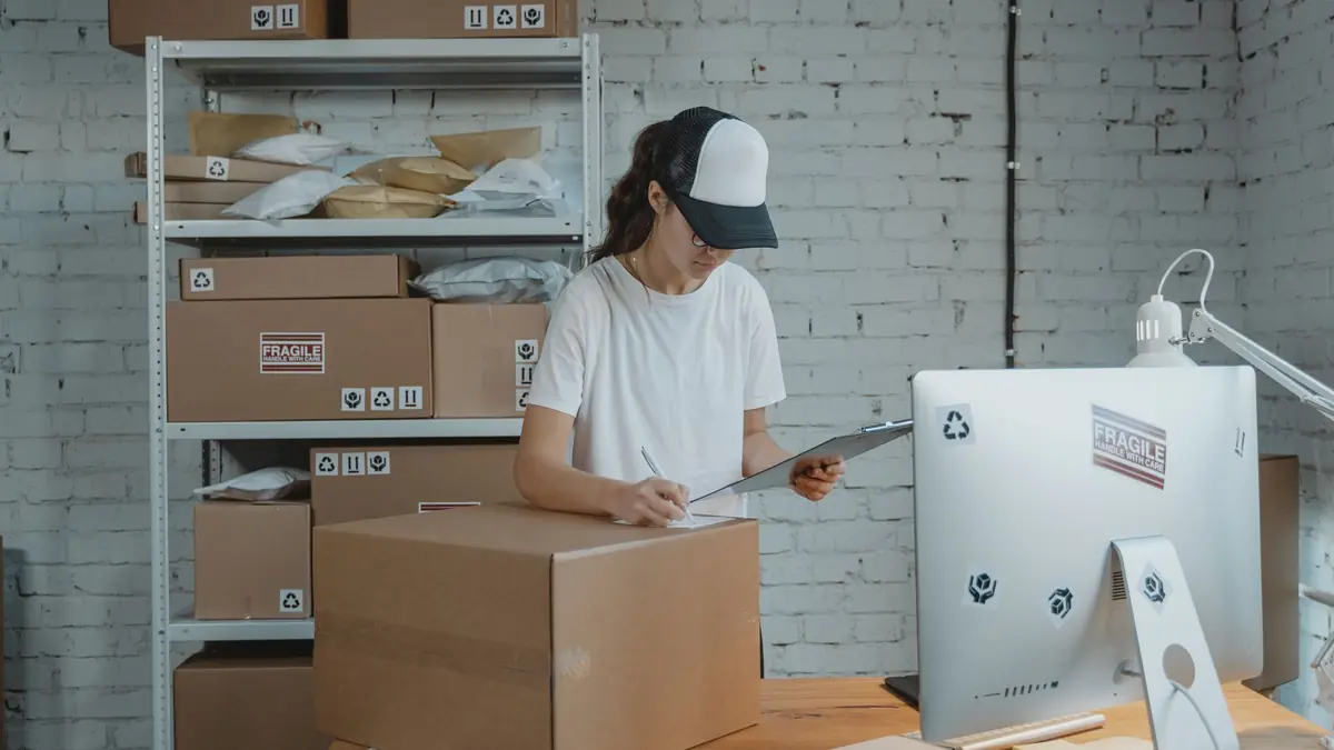 A woman in a cap writes on a clipboard beside a large cardboard box in a small packing room with shelves of parcels