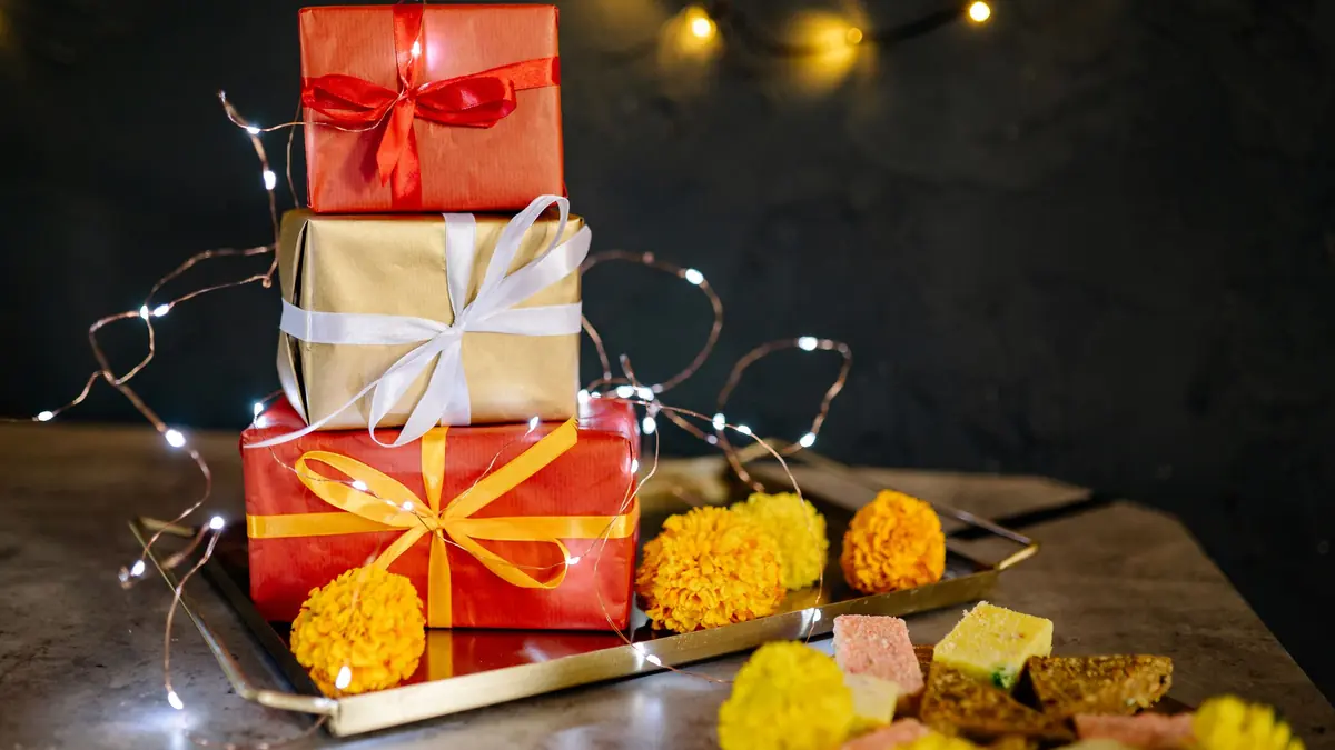 Three stacked gift boxes with ribbons on a brass tray with marigolds, sweets and fairy lights