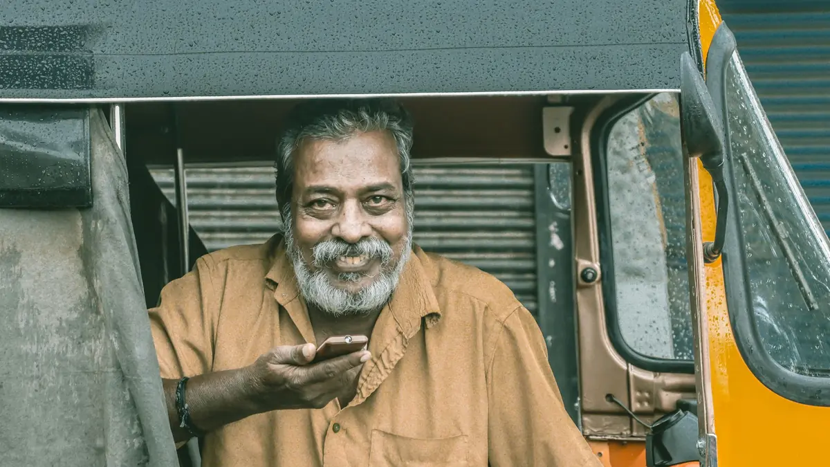 A smiling older man with a grey beard holds a smartphone while sitting in an auto-rickshaw on a rainy day
