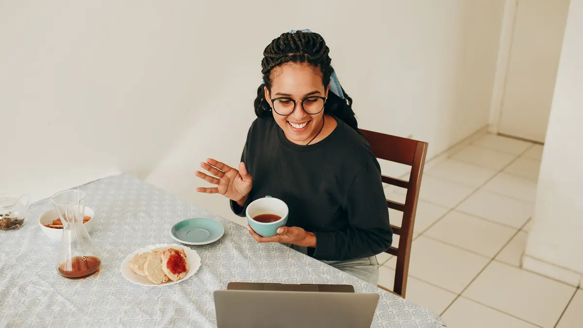 A smiling woman waves while holding a cup of tea at her dining table, with a laptop and breakfast in front of her