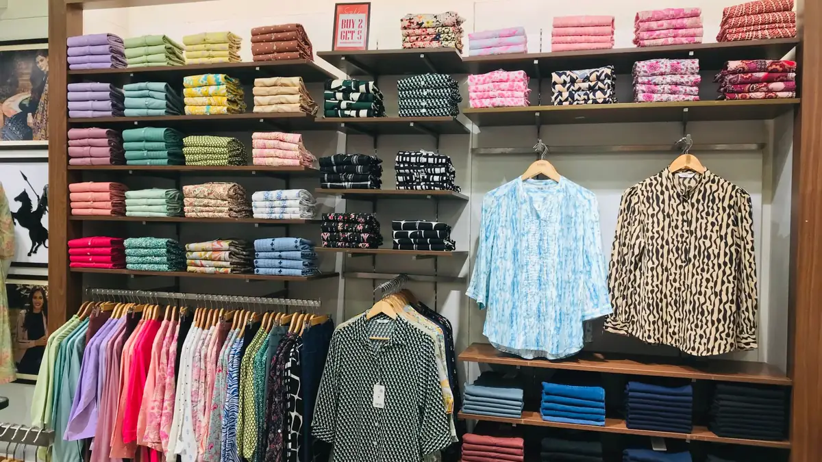 Neatly folded shirts and kurtas in many colours on wooden shelves, with printed shirts on hangers, in a clothing shop