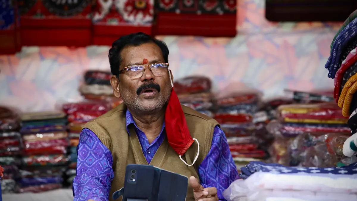 A shopkeeper holding a phone talks to someone from behind stacks of folded fabric in his clothing shop