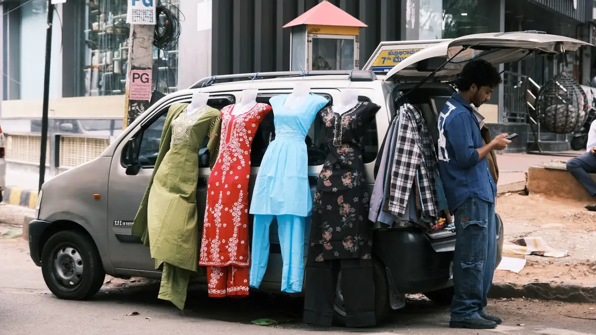 A man checks his phone beside a parked car hung with kurtas and salwar suits for sale on a Bengaluru street