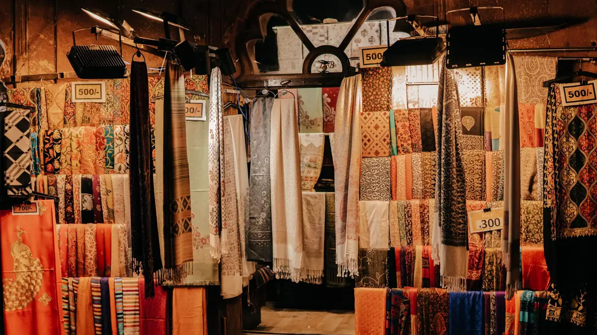 Shawls and stoles in rich colours hang in a market shop, with handwritten price cards reading 500 and 300 rupees