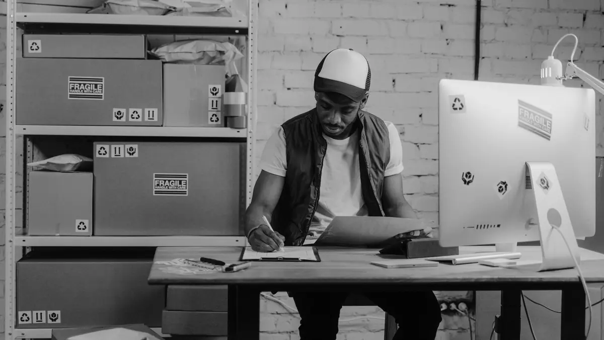 A man in a cap fills in paperwork on a clipboard at a desk in a stockroom, with labelled parcels on shelves beside him, in black and white