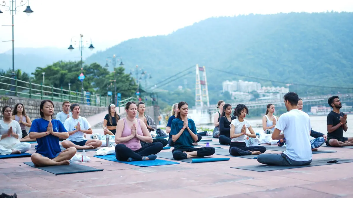 A group sits cross-legged on yoga mats with palms joined, led by an instructor, on a riverside promenade in Rishikesh with green hills behind