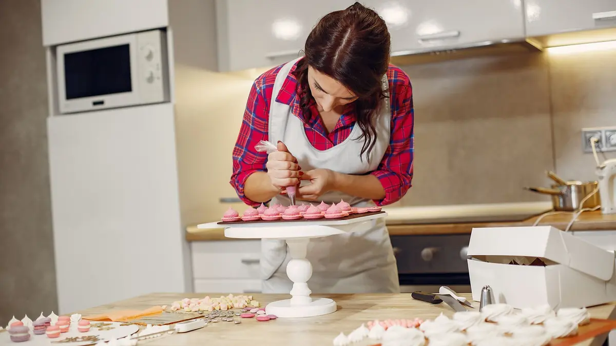 A woman in an apron pipes pink cream onto rows of small sweets on a cake stand in a home kitchen, with white cake boxes on the counter