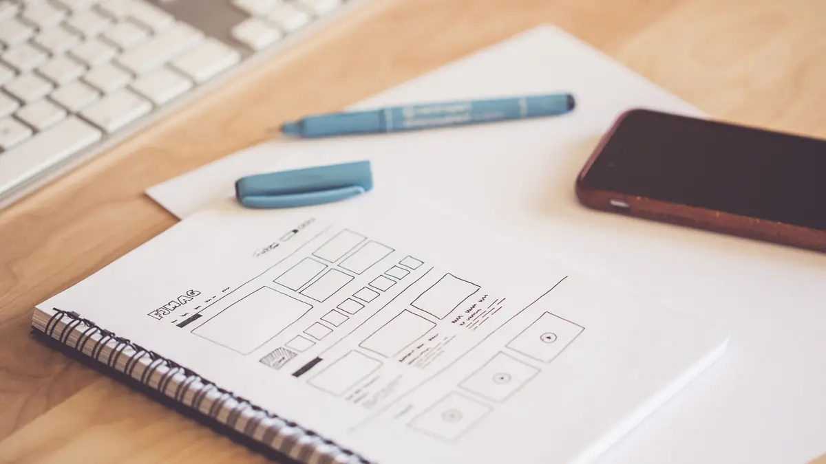 A notebook with a hand-drawn web page wireframe lies beside a smartphone, pens and a keyboard on a wooden desk
