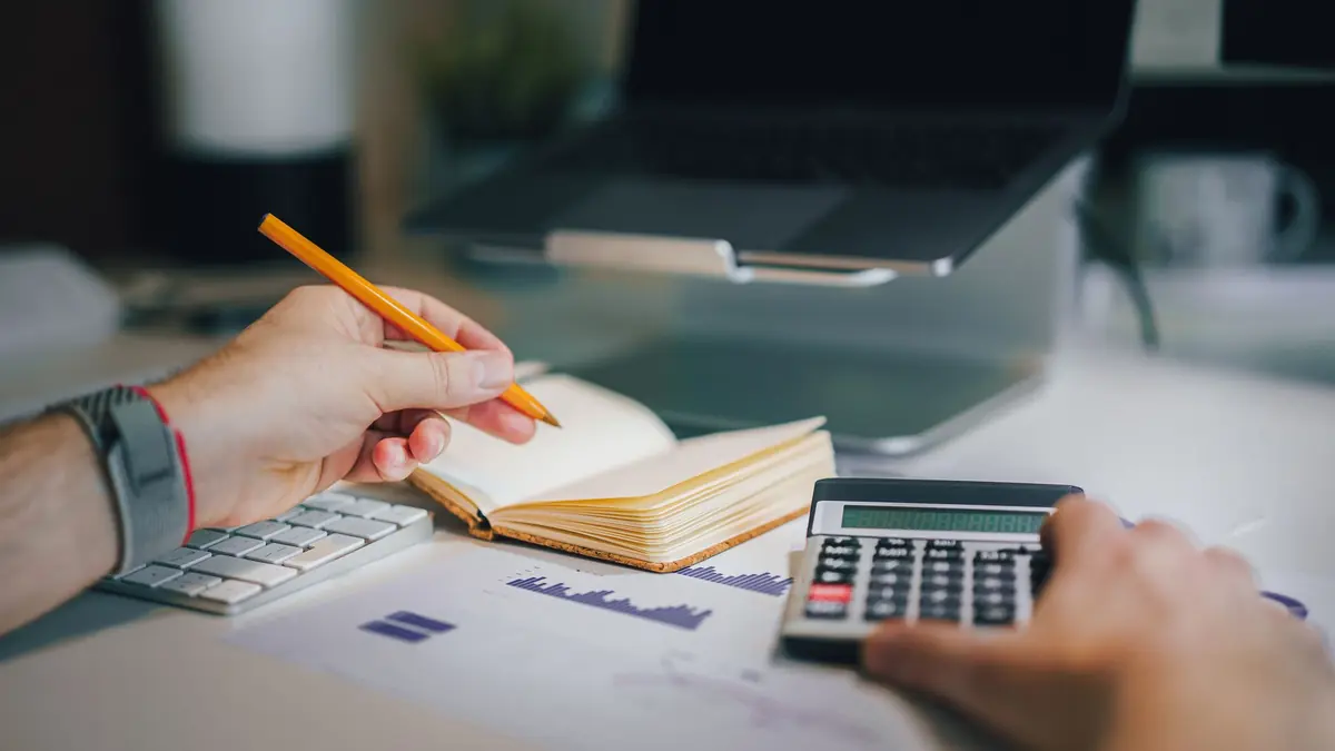 Hands holding a pencil over an open notebook and working a calculator on a desk with printed charts and a laptop