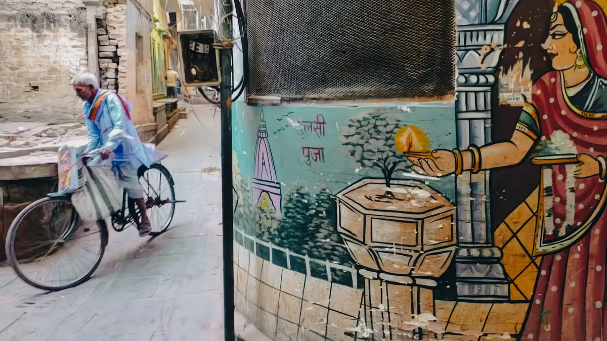A man cycles with bags hanging from his handlebars through a narrow lane in Varanasi, past a painted wall mural