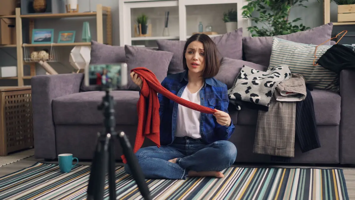 A woman sitting on a rug holds up a red knitted garment while filming herself with a phone on a tripod in her living room