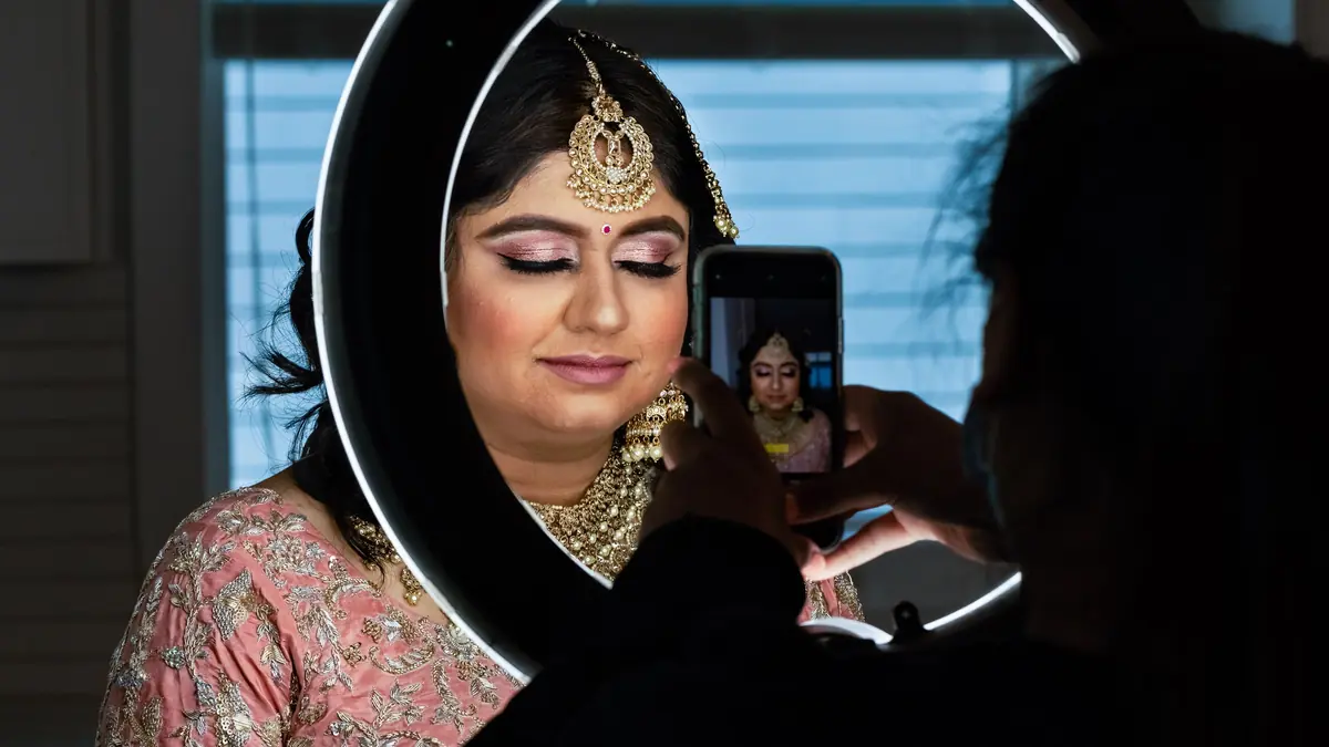 A bride in gold jewellery and a pink lehenga is photographed on a phone through a ring light
