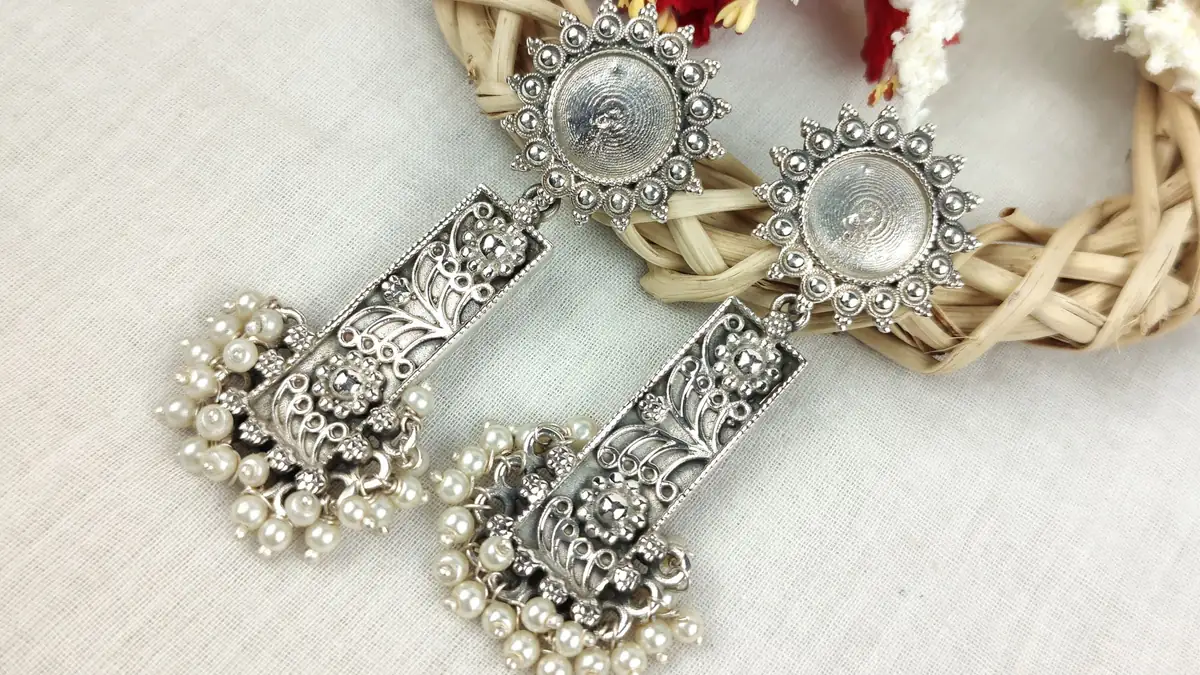 A pair of oxidised silver earrings with pearl drops laid on a white cloth beside a wicker ring