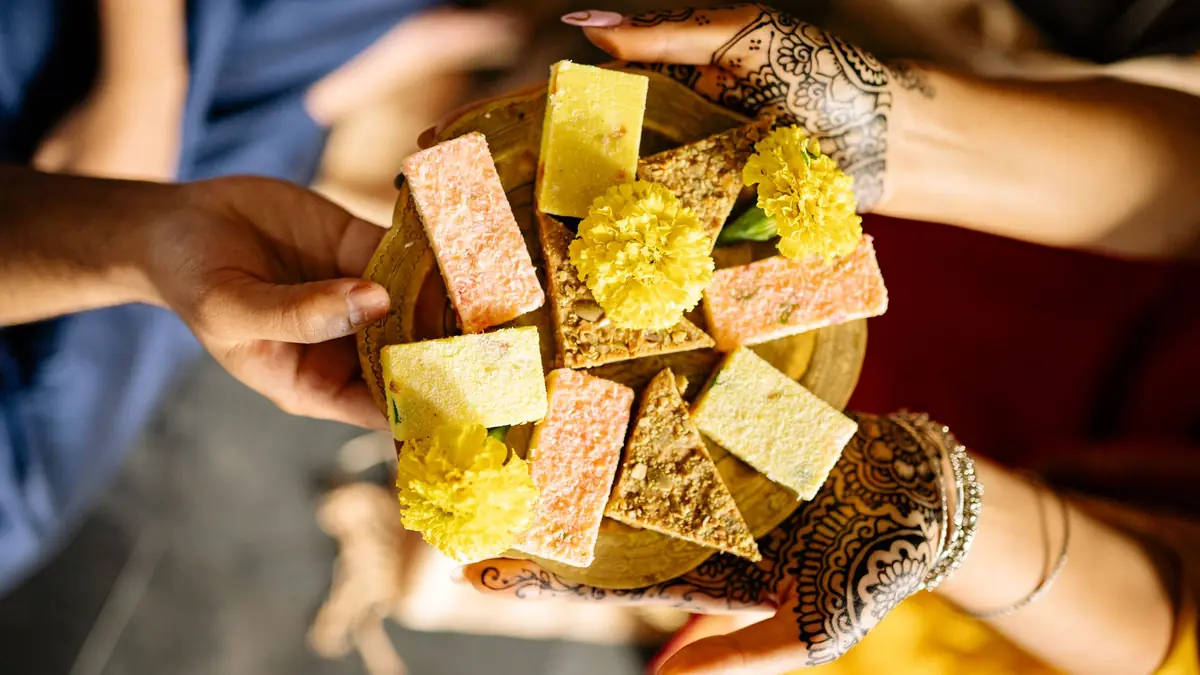 Two hands, one decorated with henna, pass a plate of Indian sweets and marigold flowers