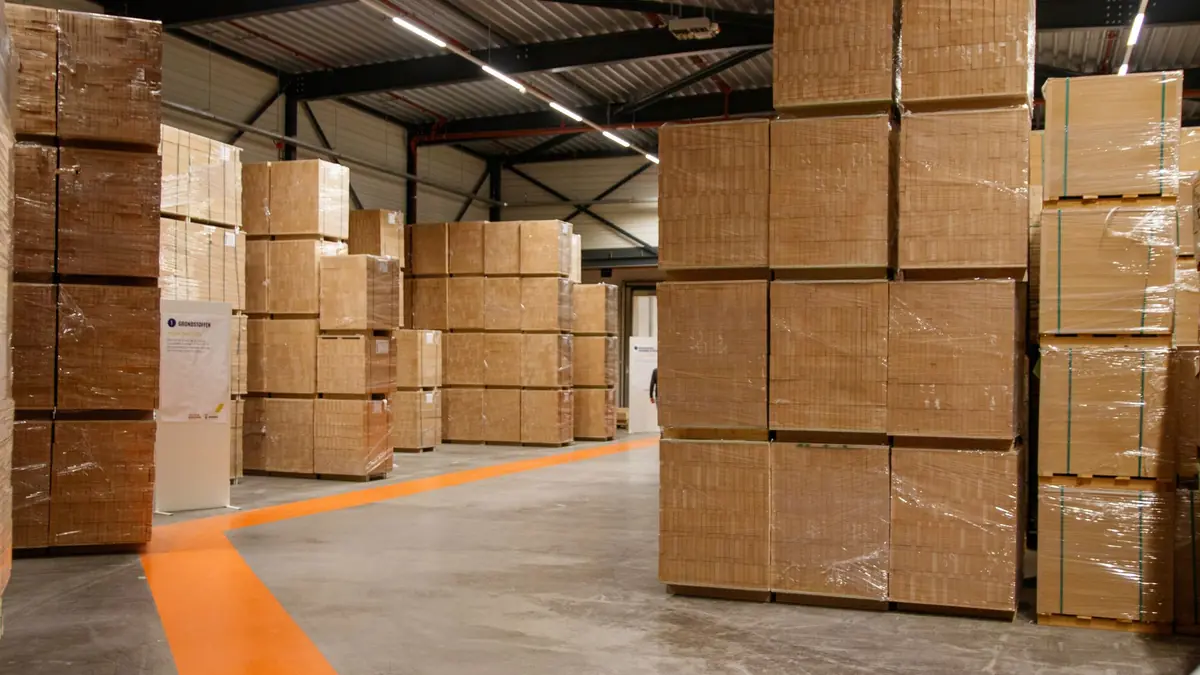 Tall stacks of shrink-wrapped cardboard cartons on pallets in a warehouse with an orange line painted on the floor