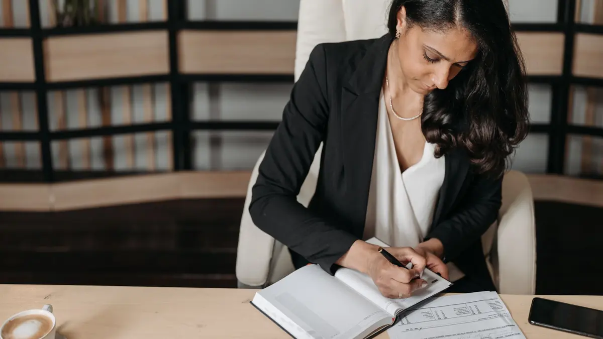 A woman in a black blazer writes in a notebook at a desk beside a cup of coffee, papers and a phone