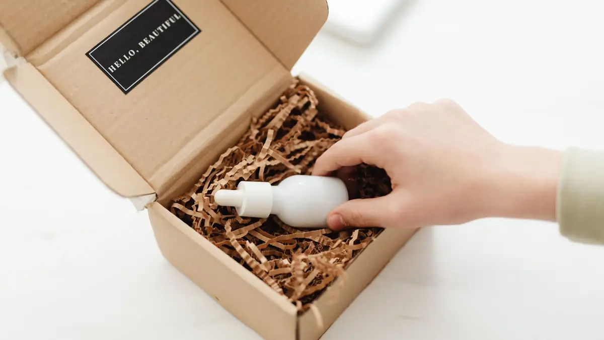A hand lifts a white dropper bottle out of a cardboard box filled with shredded paper packing