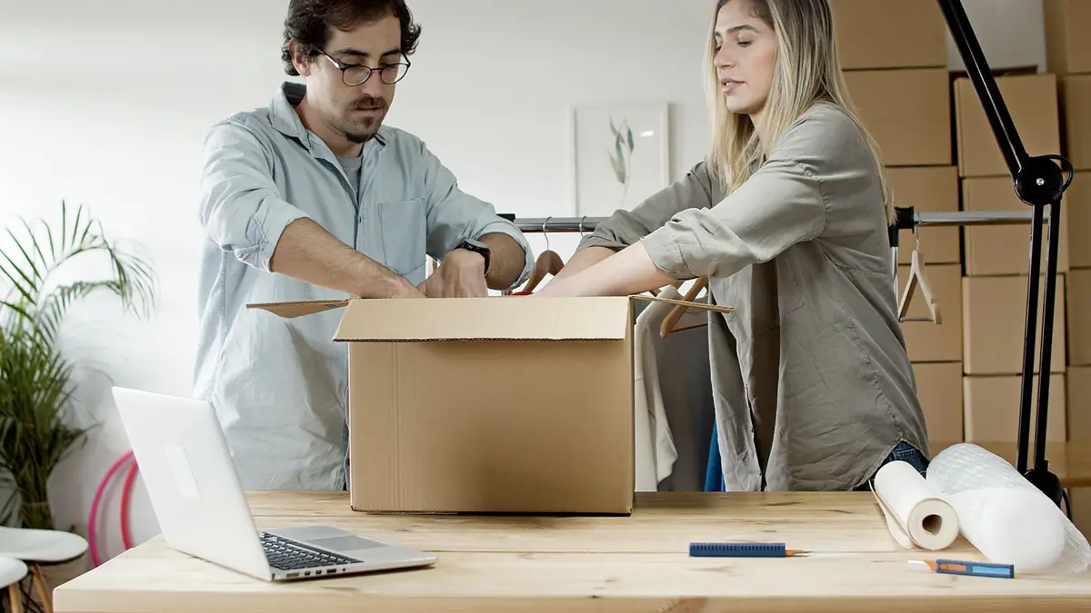 A man and a woman pack clothes into a large cardboard box on a table, with an open laptop beside them and shipping boxes stacked behind