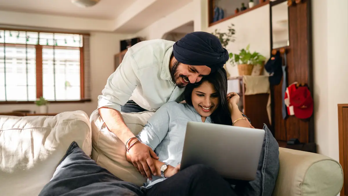 A smiling couple share a laptop on a sofa in their living room