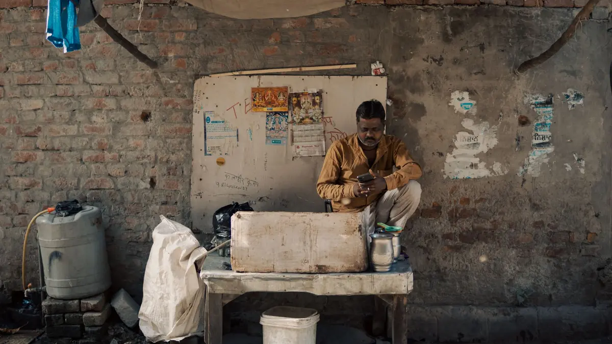 A street vendor sits behind his stall against a brick wall, looking at his mobile phone