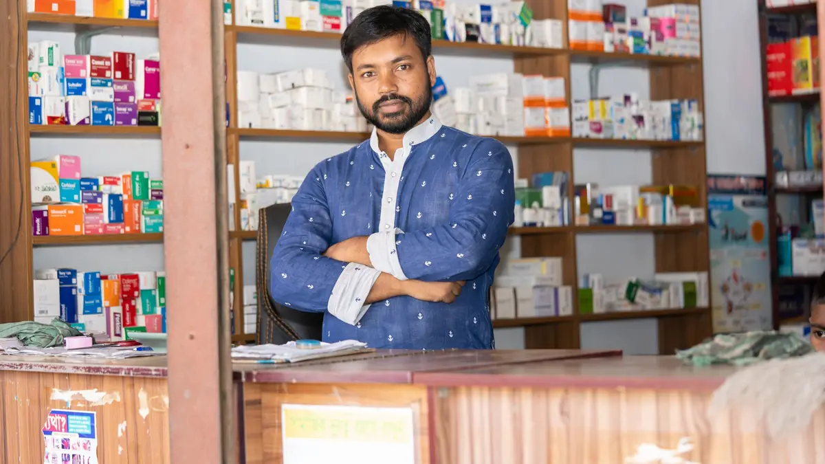 A pharmacist stands with folded arms behind the counter of a small chemist shop lined with shelves of medicine boxes