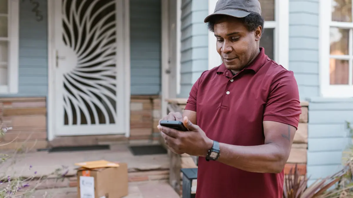 A man in a maroon polo shirt checks his phone outside a house, with a delivered parcel on the porch steps