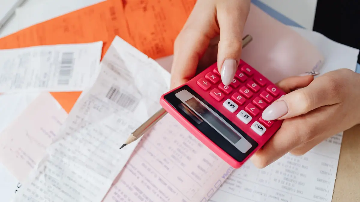 Hands with a pink calculator over a pile of paper receipts and bills on a desk