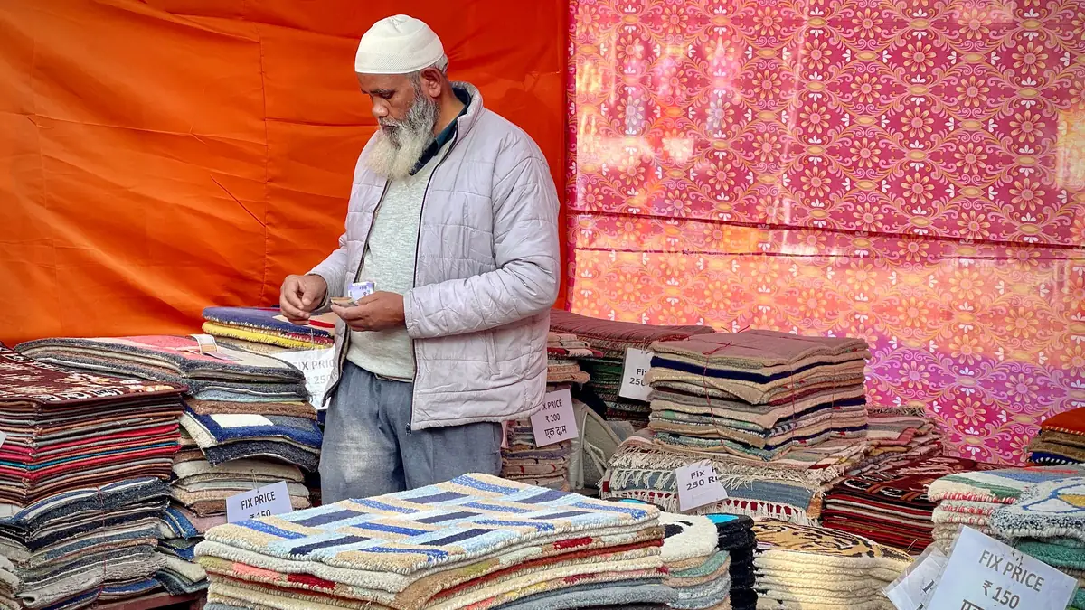 A vendor counts money beside stacks of rugs marked with handwritten fixed-price tags at a market stall