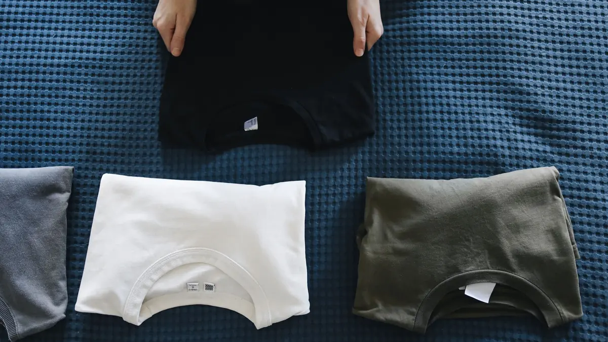 Hands lay out folded black, white and grey T-shirts on a blue bedspread