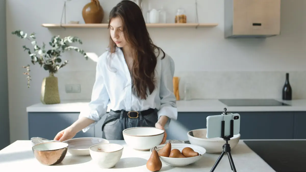 A smartphone on a small tripod records a woman arranging ceramic bowls on a kitchen counter