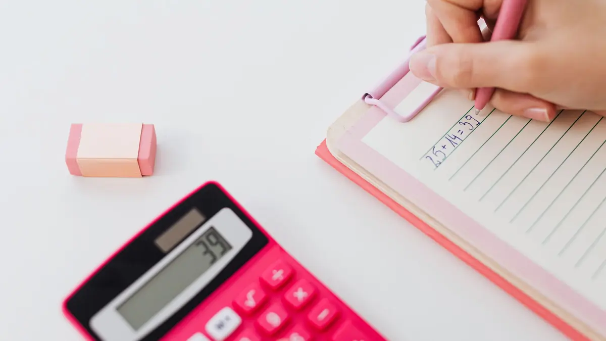 A hand writes on a clipboard notepad beside a pink calculator and an eraser