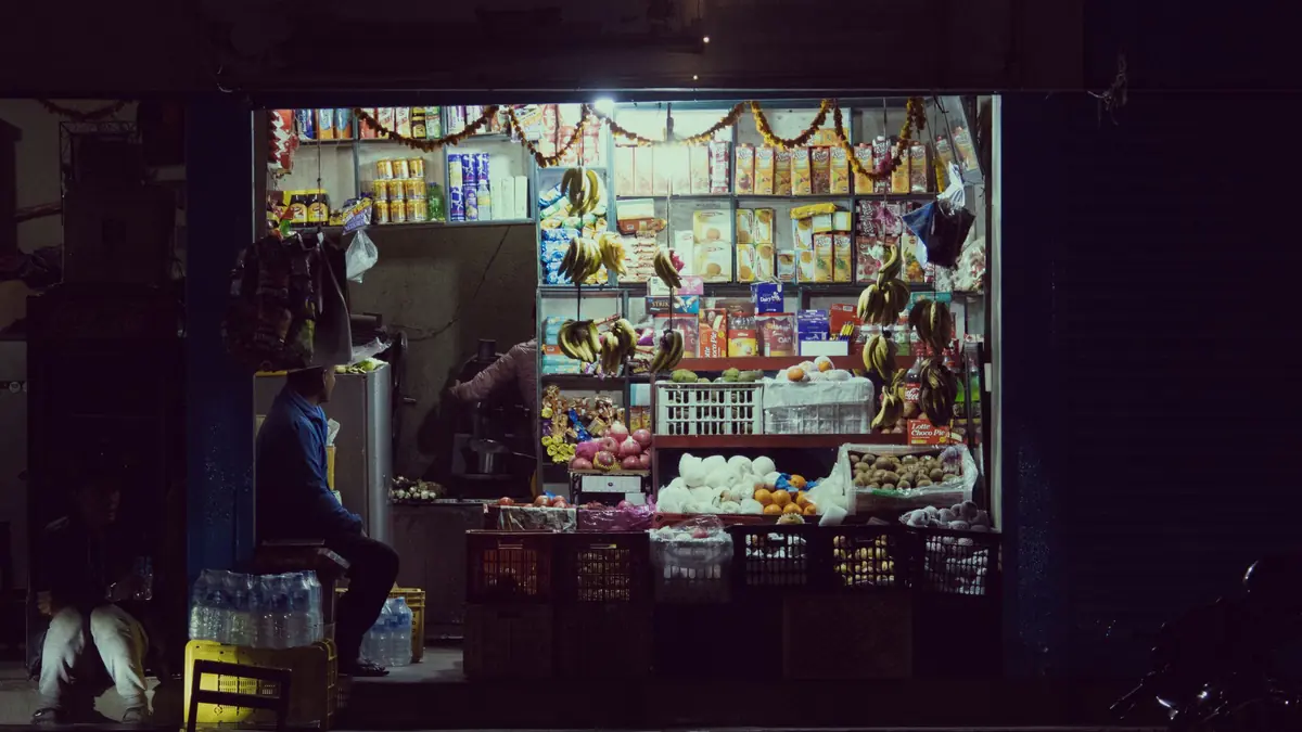 A small grocery shop glows at night, its shelves stocked with packets, bananas and eggs, a shopkeeper seated at the front