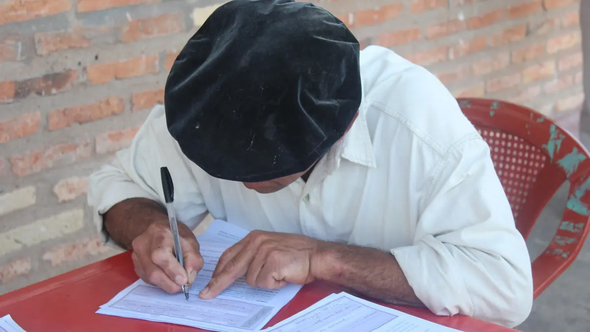 A man in a cap leans over a red table to fill in a printed form with a pen