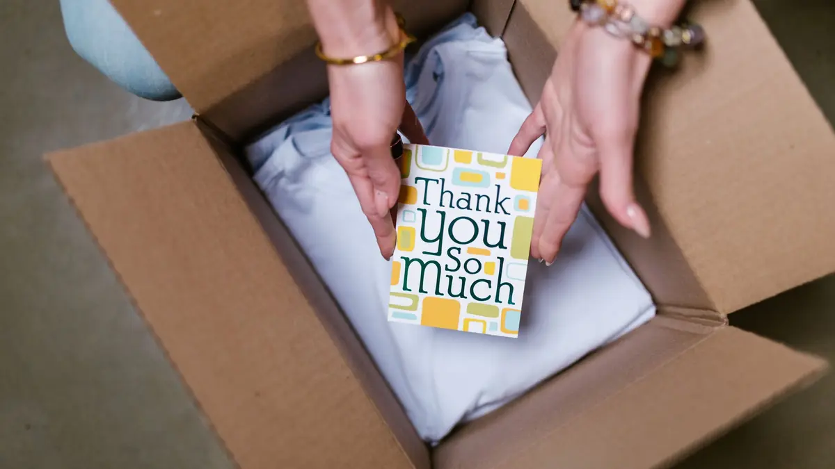 Hands place a thank-you card on folded clothing in an open cardboard box