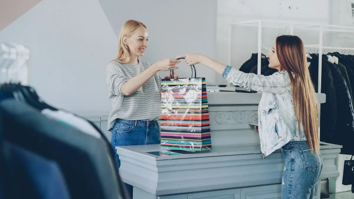 A smiling shop assistant hands a striped shopping bag across the counter to a customer in a clothing store