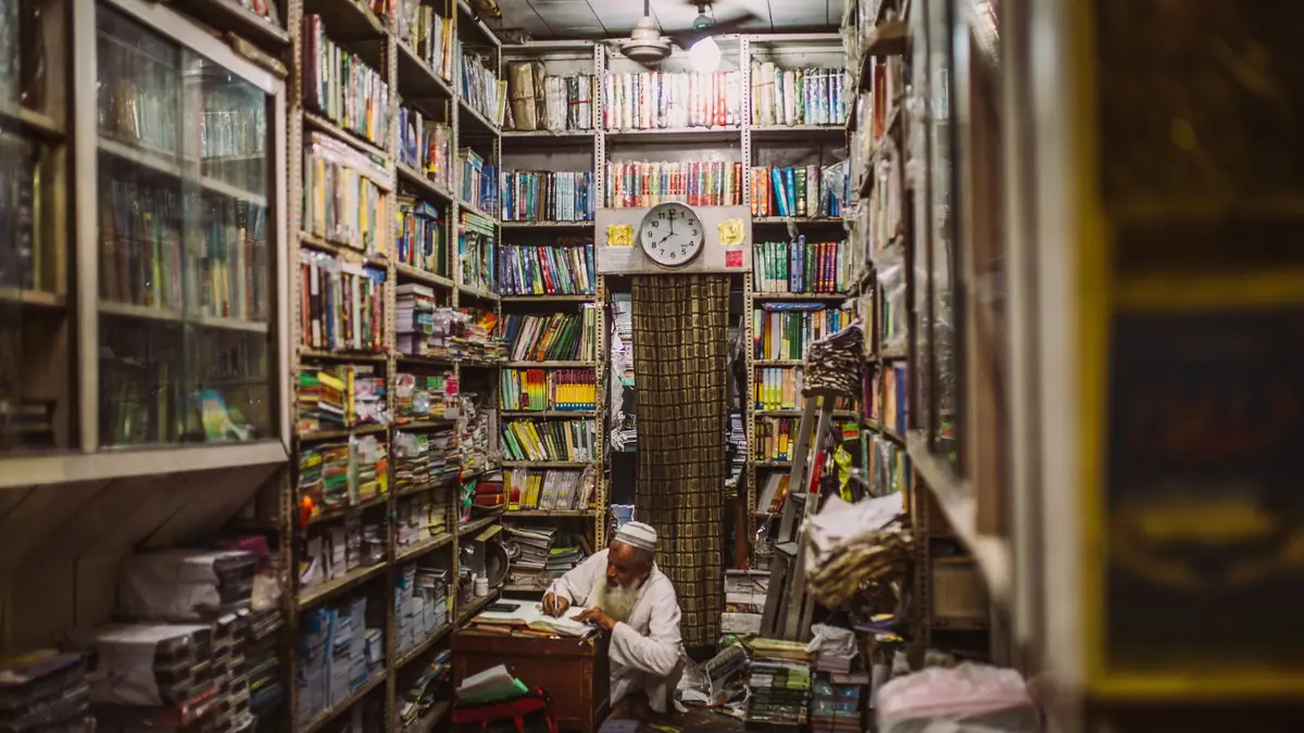 An elderly bookseller reads at his desk at the end of a narrow Delhi bookshop lined floor to ceiling with books