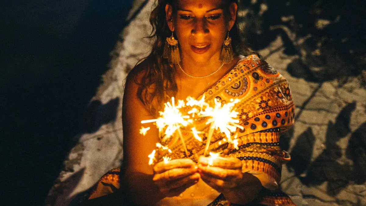 A woman in a sari holds lit sparklers close to her face at night, their glow lighting the scene