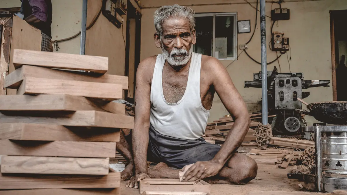 A senior carpenter sits among stacked wooden planks in a traditional workshop, smoothing a piece of wood