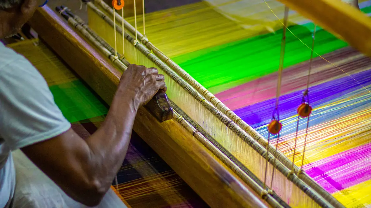 A weaver's hand works the wooden beam of a handloom strung with bright green, yellow and purple threads