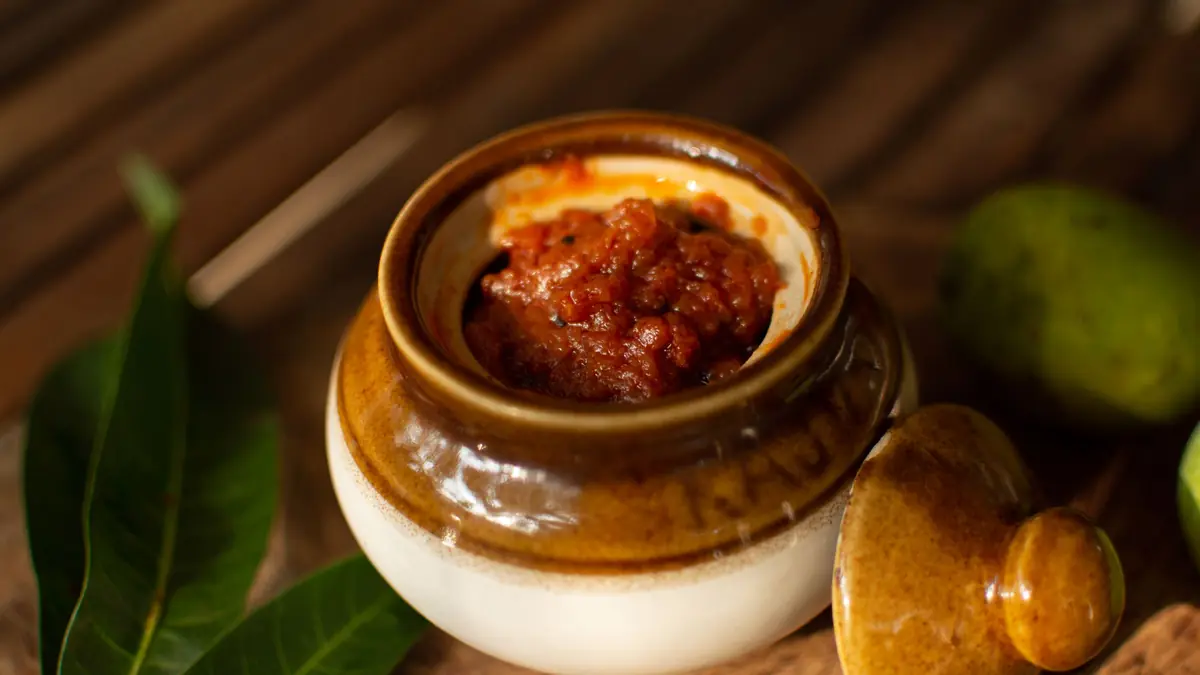A ceramic jar of red mango pickle beside green mangoes and mango leaves