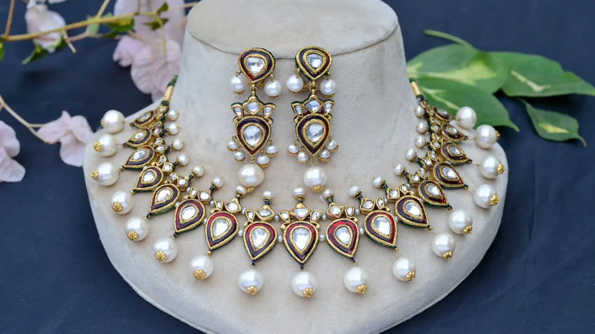 A kundan necklace and earring set with pearl drops displayed on a cream velvet bust, with pink flowers and green leaves around it