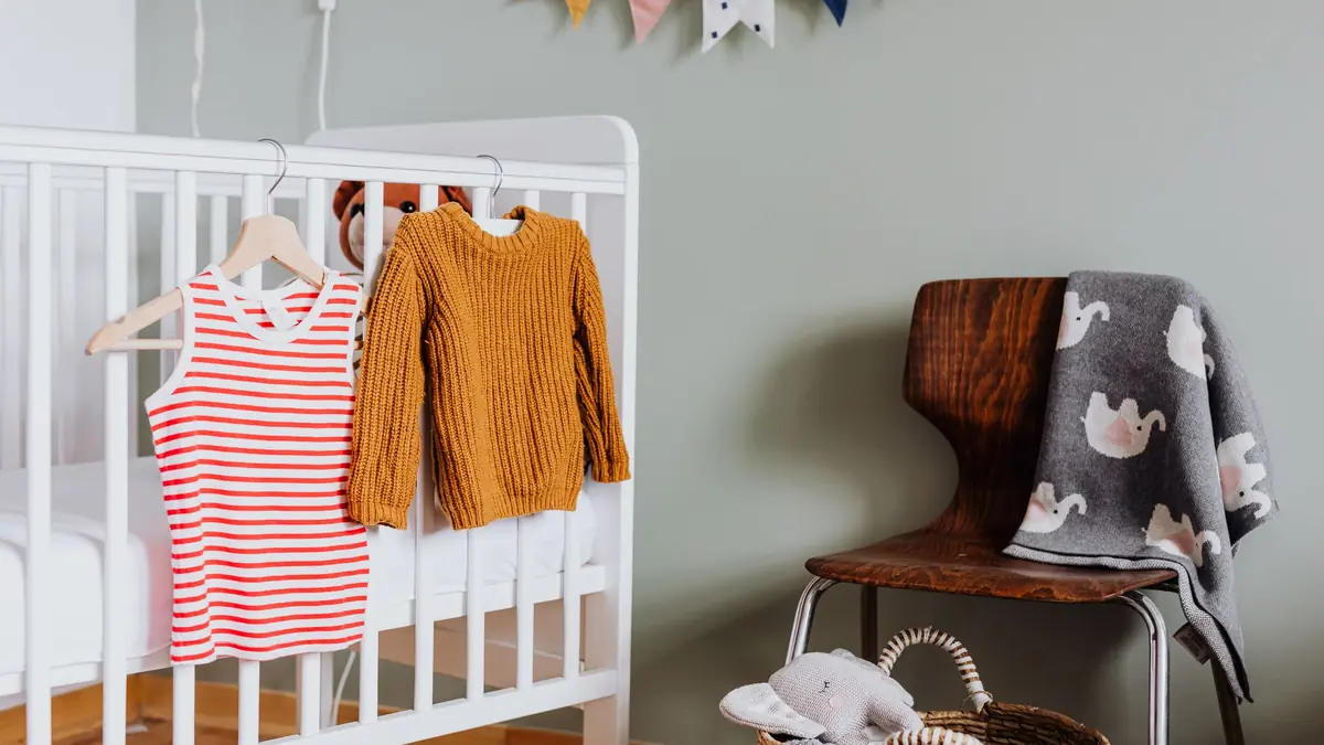 A striped child's top and a mustard knitted sweater hang on the side of a white cot in a nursery