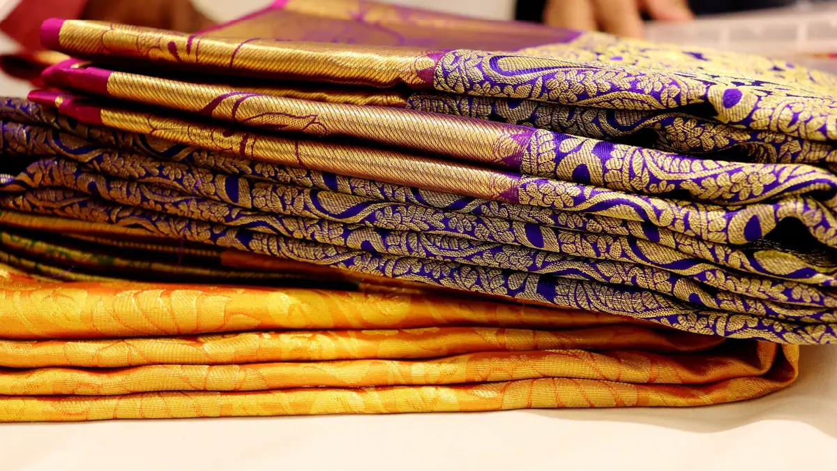A stack of folded Kanchipuram silk sarees in purple and gold with zari patterns
